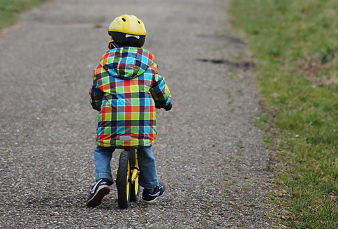 Man sieh ein Kind auf einem Laufrad, von hinten. Rechts und links des Weges sieht man Rasen.