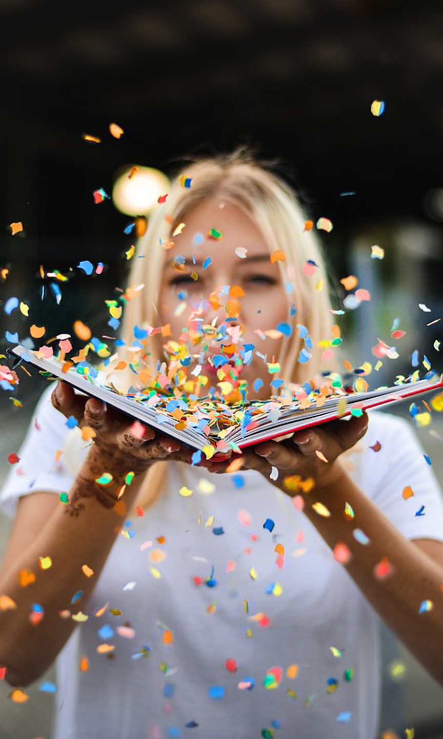 Eine Frau hat ein offenes Buch in den Händen und pustet Konfetti durch die Luft.