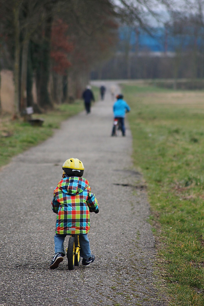 Man sieh ein Kind auf einem Laufrad, von hinten. Rechts und links des Weges sieht man Rasen.