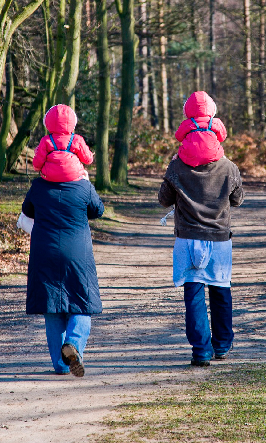 Eine Familie von hinten im Wald. Mutter und Vater tragen die Kleinkinder auf den Schultern.