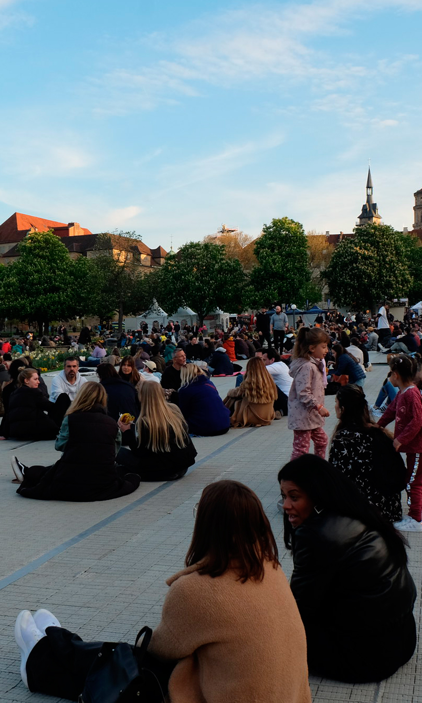Im Vordergrund sitzen Menschen auf dem Boden. In Hintergrund sieht man Stuttgarter Gebäude.