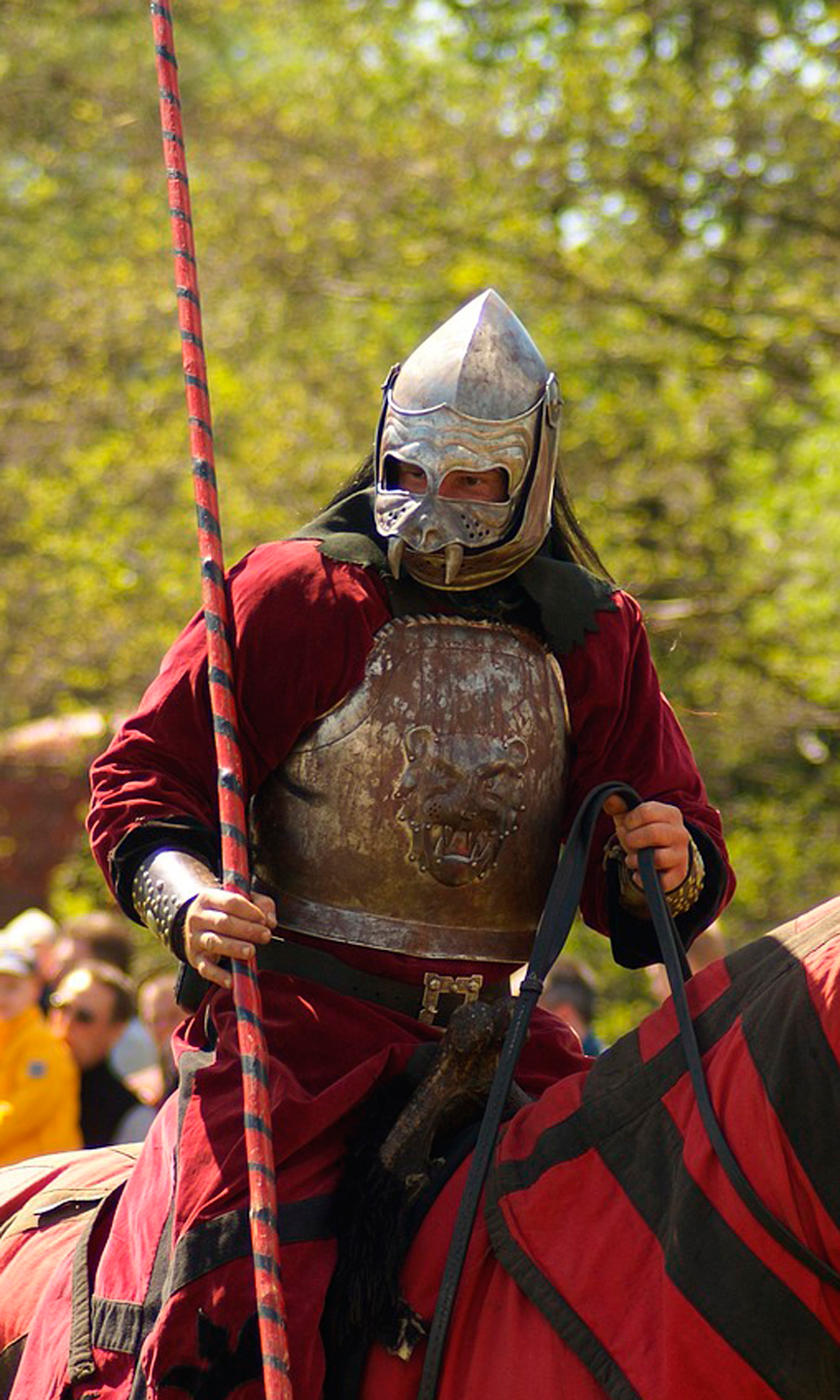 Ein Ritter sitzt mit Rüstung, Helm und Lanze auf einem Pferd.