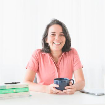 Frau sitzt an einem Tisch mit einer Tasse in der Hand und lächelt.