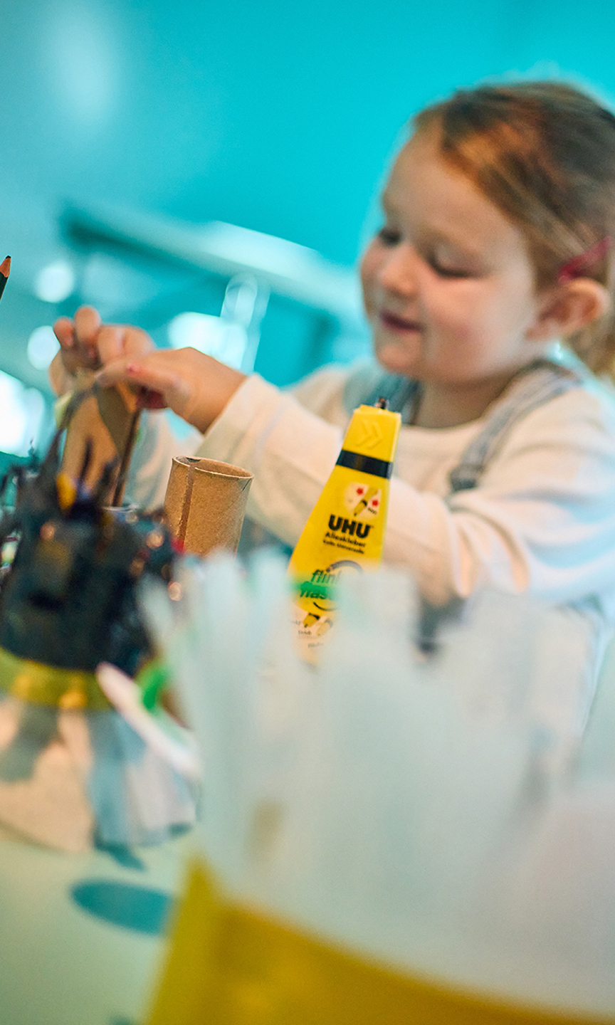 Mädchen sitzt an einem Basteltisch. Vor ihr liegen Bastelmaterialien.