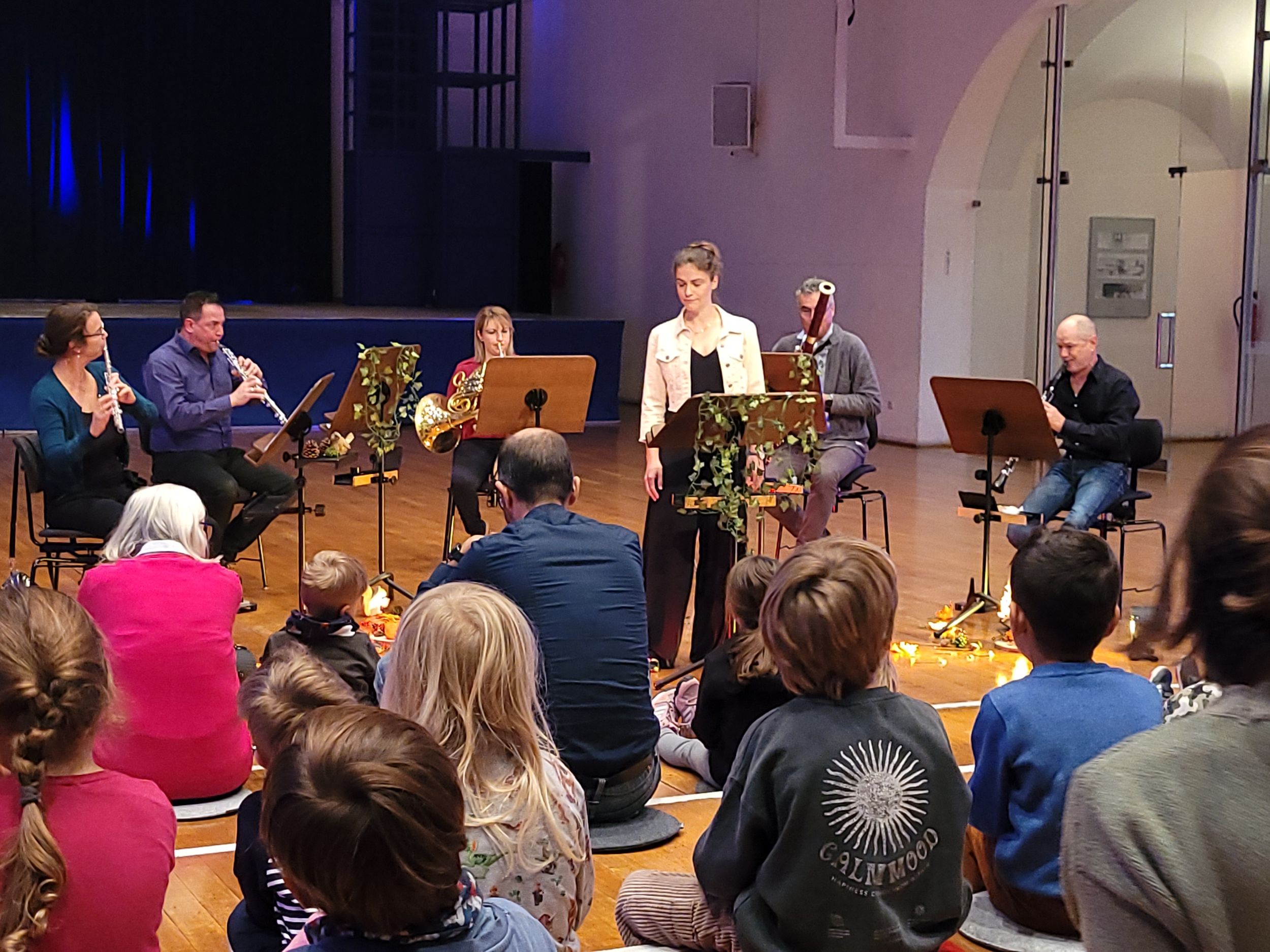 Eine Gruppe von Erwachsenen und Kinder sitzt auf dem Boden vor einem Orchester.