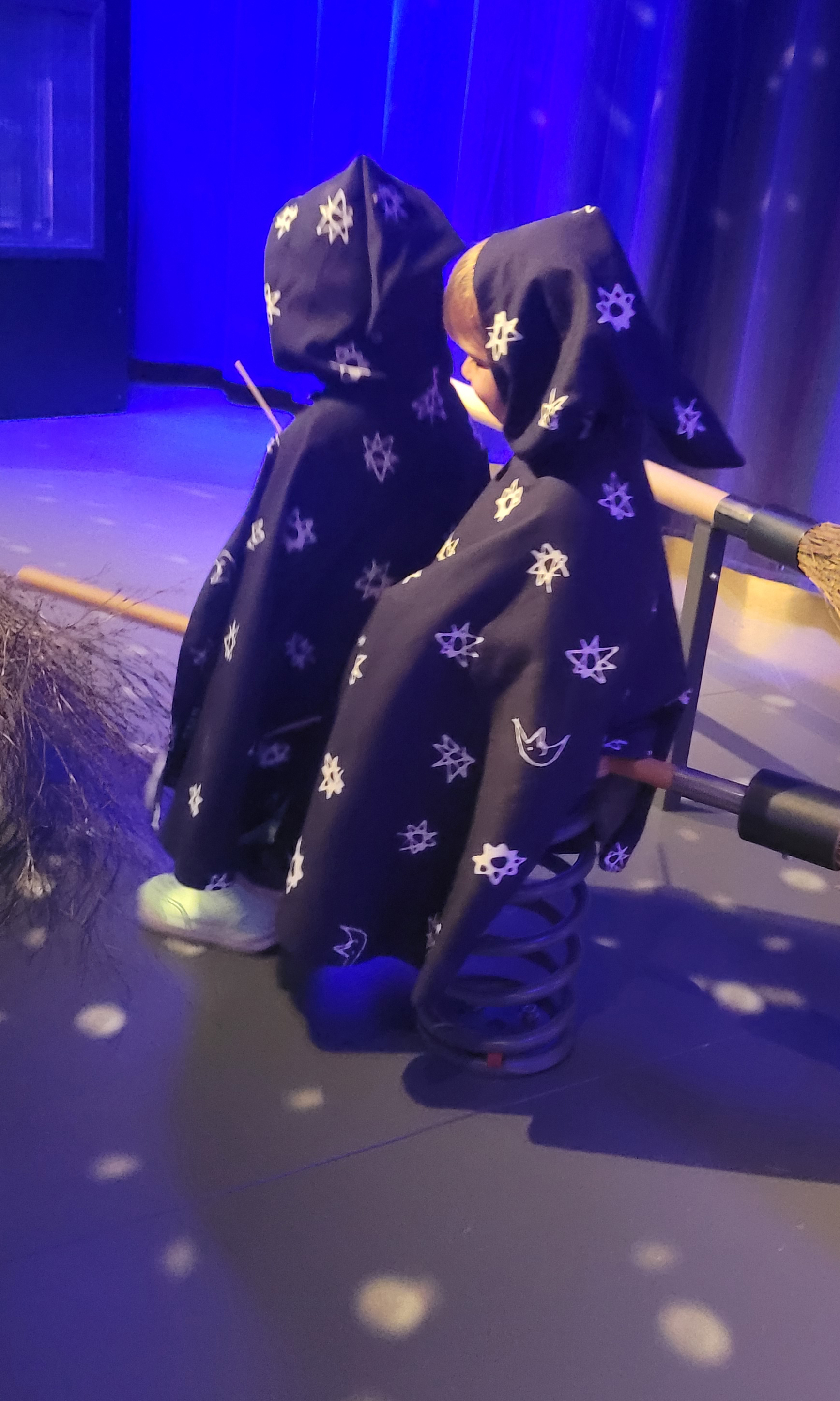 Zwei kleine Kinder haben Zaubermäntel an und sitzen gemeinsam auf einem Hexenbesen.