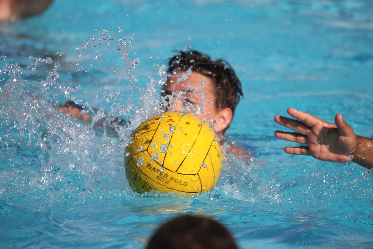 Ein Ball platscht ins Wasser, im Hintergrund Wasser-Polo-Spieler.