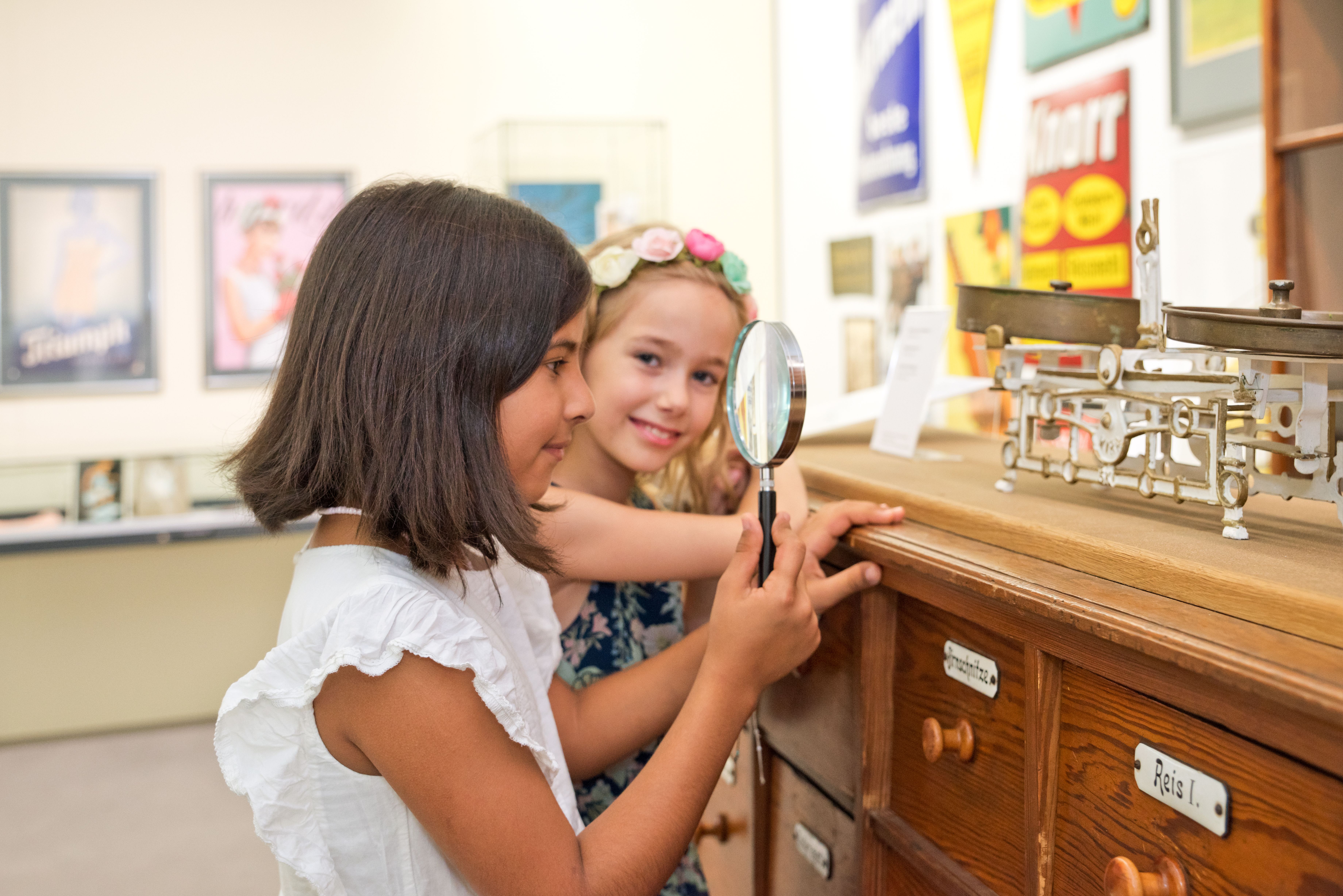 Ein Mädchen untersucht mit einer Lupe ein altes Gerät, daneben ein Mädchen im Hintergrund am lächeln