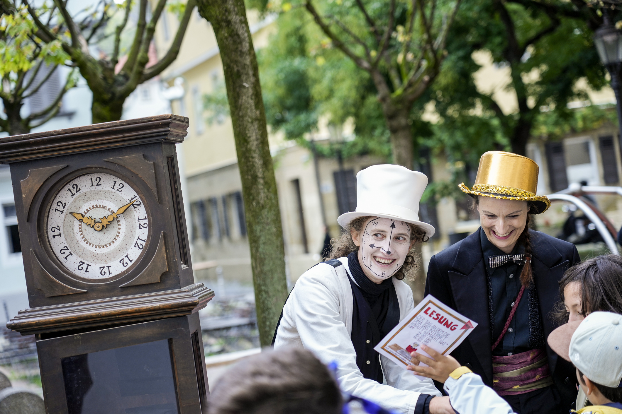 Links eine große Standuhr, rechts Schuaspielende mit aufgemalter Uhr im Gesicht.