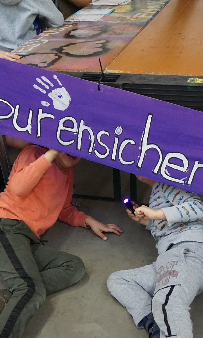 Zwei Kinder liegen mit einer Taschenlampe unter einem Tisch. Plakat Aufschrift "Spurensicherung"