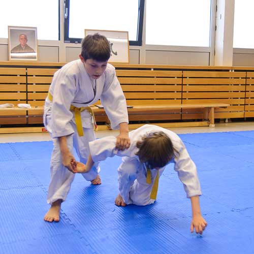 Zwei Jungen im Aikido-Anzug kämpfen: Einer kniet am Boden, einer steht darüber und hält ihn am Arm.