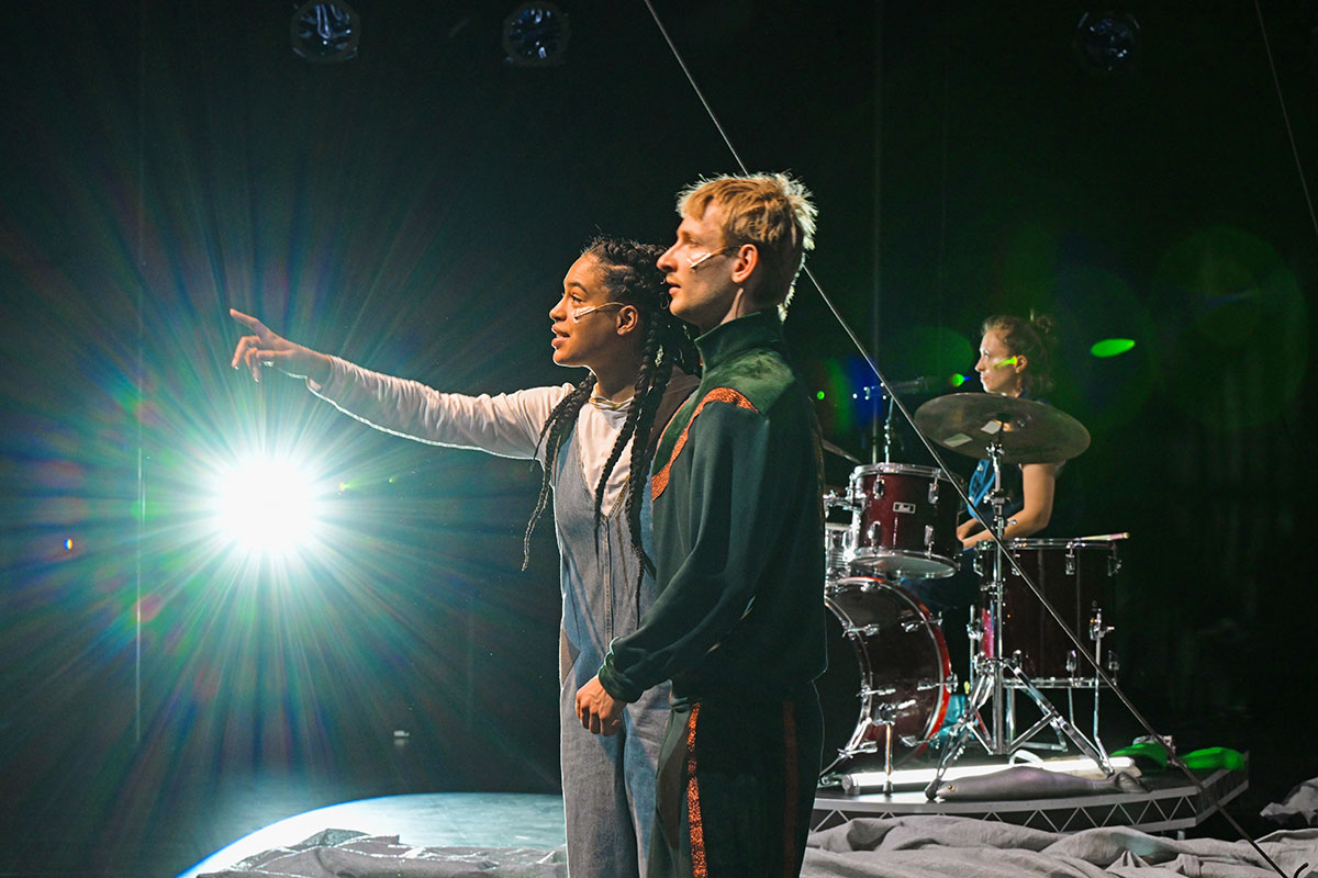 Zwei Schauspieler:innen stehen auf einer Bühne und zeigen ins Publikum