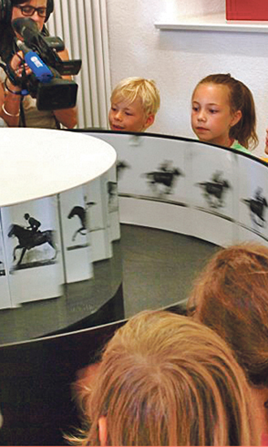 Kinder stehen um ein Museumsobjekt herum. Das Objekt zeigt eine Art Daumenkino.