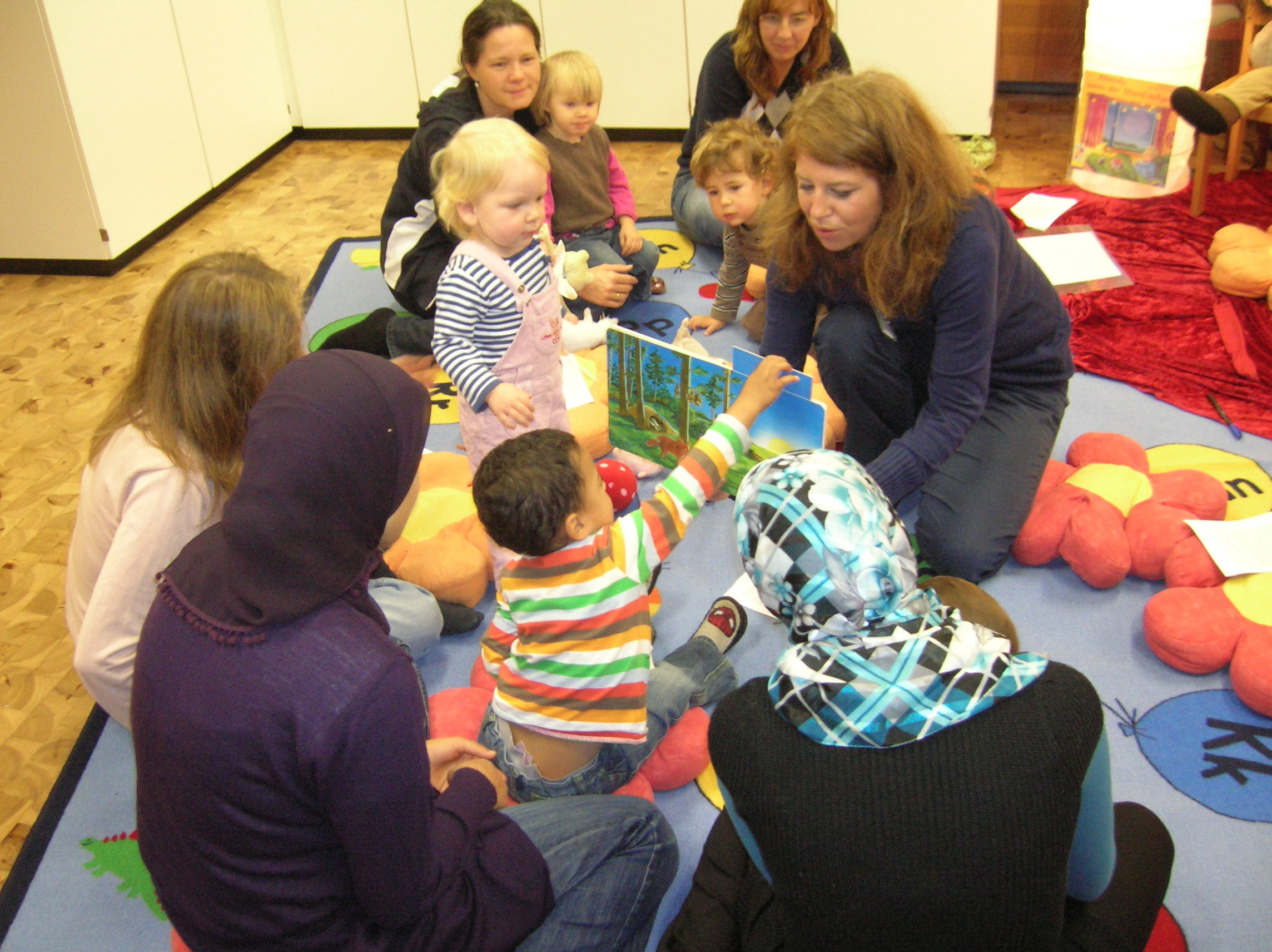 Junge Mütter und Kleinkinder sitzen auf dem Boden um eine Frau mit Bilderbuch herum