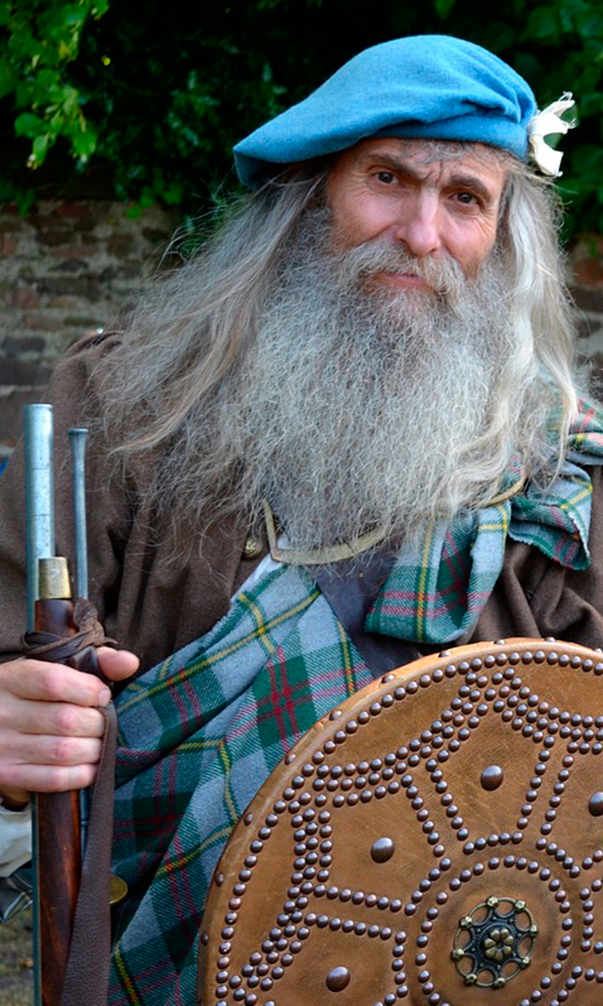 Zeigt einen Mann mit langen weißen Haaren und Bart, der mittelalterliche Kleidung trägt.