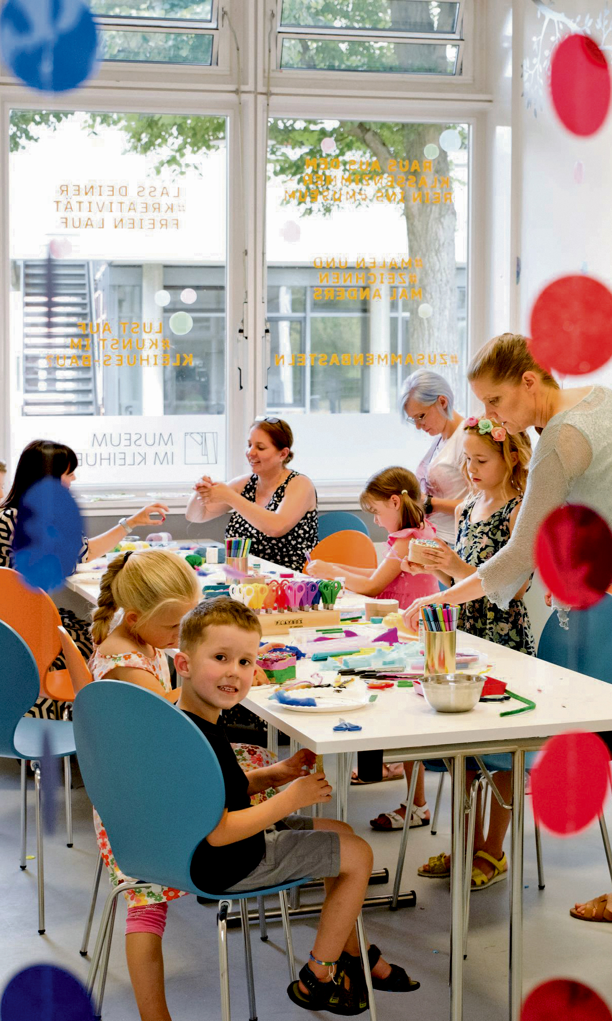 Kinder sitzen an einem Tisch verteilt und Basteln, eine Frau erklärt etwas.