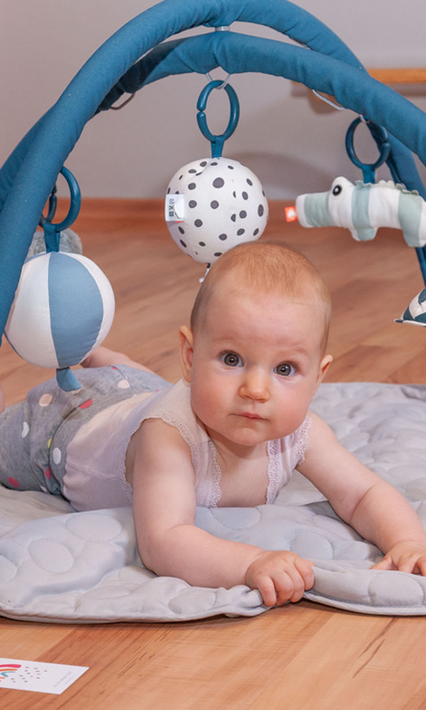 Ein Säugling liegt auf dem Bauch unter einem Spielbogen.