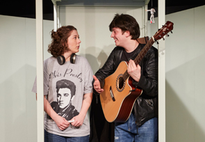 Zwei Personen stehen in einer Telefonzelle. Der Mann hält eine Gitarre in den Händen.