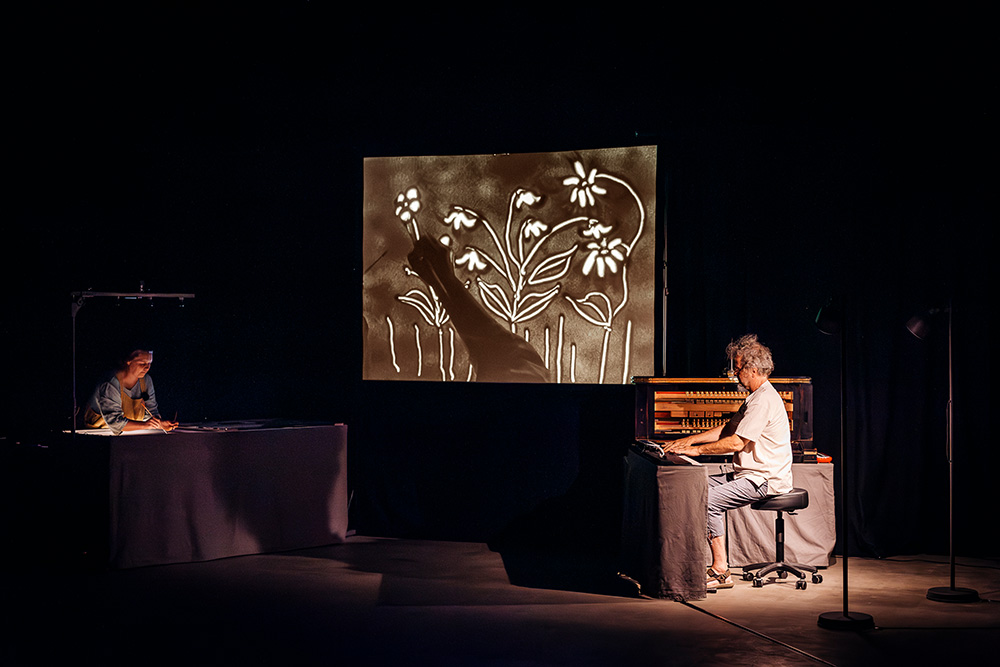 Die Künslterin Sandrine malt ein Sandbild und wird vom Klavier begleitet.