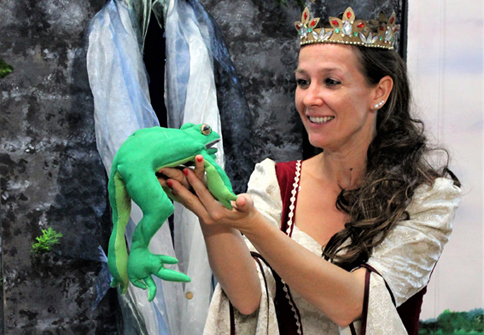 Dornröschen steht vor einem Schloss und schaut einen Frosch in ihren Händen an.