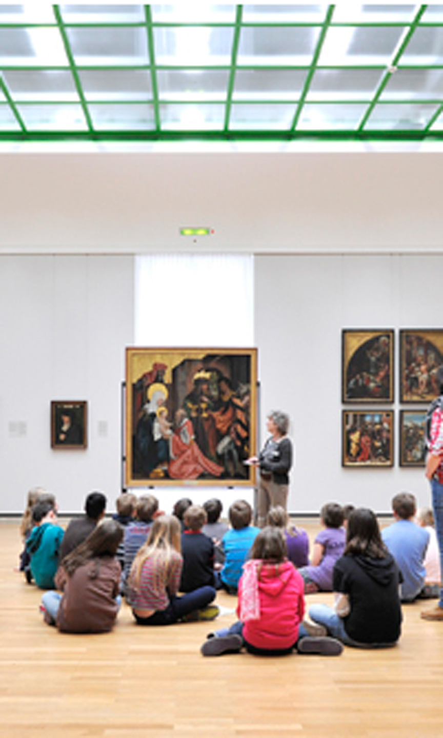 Eine Gruppe von Kinder und Jugendlichen sitzt vor einem Gemälde auf dem Boden.