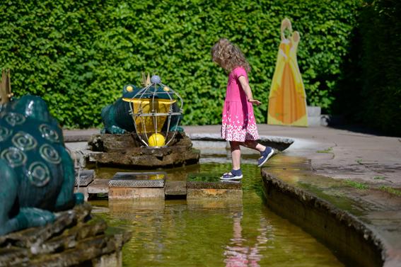 Mädchen läuft über Steine in einem Wasserbecken. Daneben steht einen Figur des Froschkönigs.