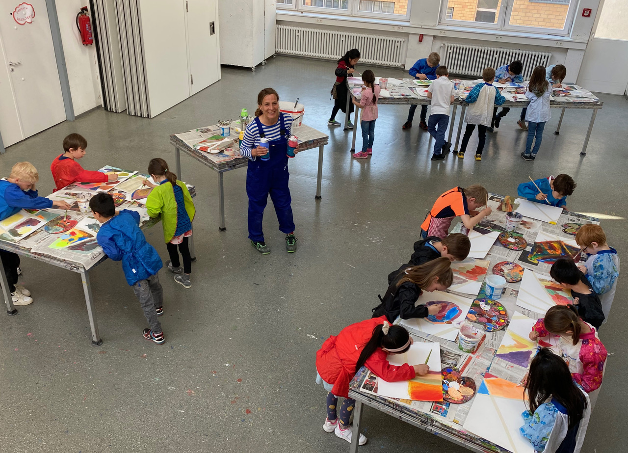 Pädagogin mit vielen Kindern in einem Raum. Die Kinder stehen in Gruppen an Tischen und malen.