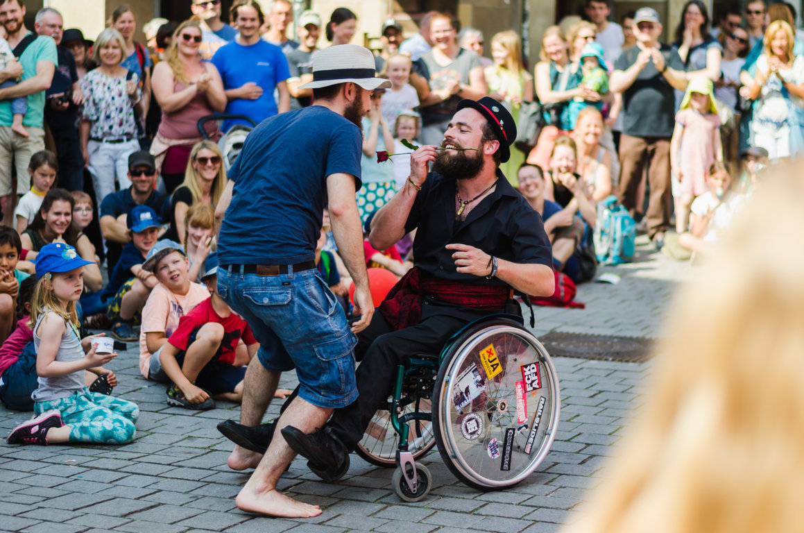 Zwei Männer mit Hut tanzen. Einer sitzt im Rollstuhl und hat eine Blume im Mund.