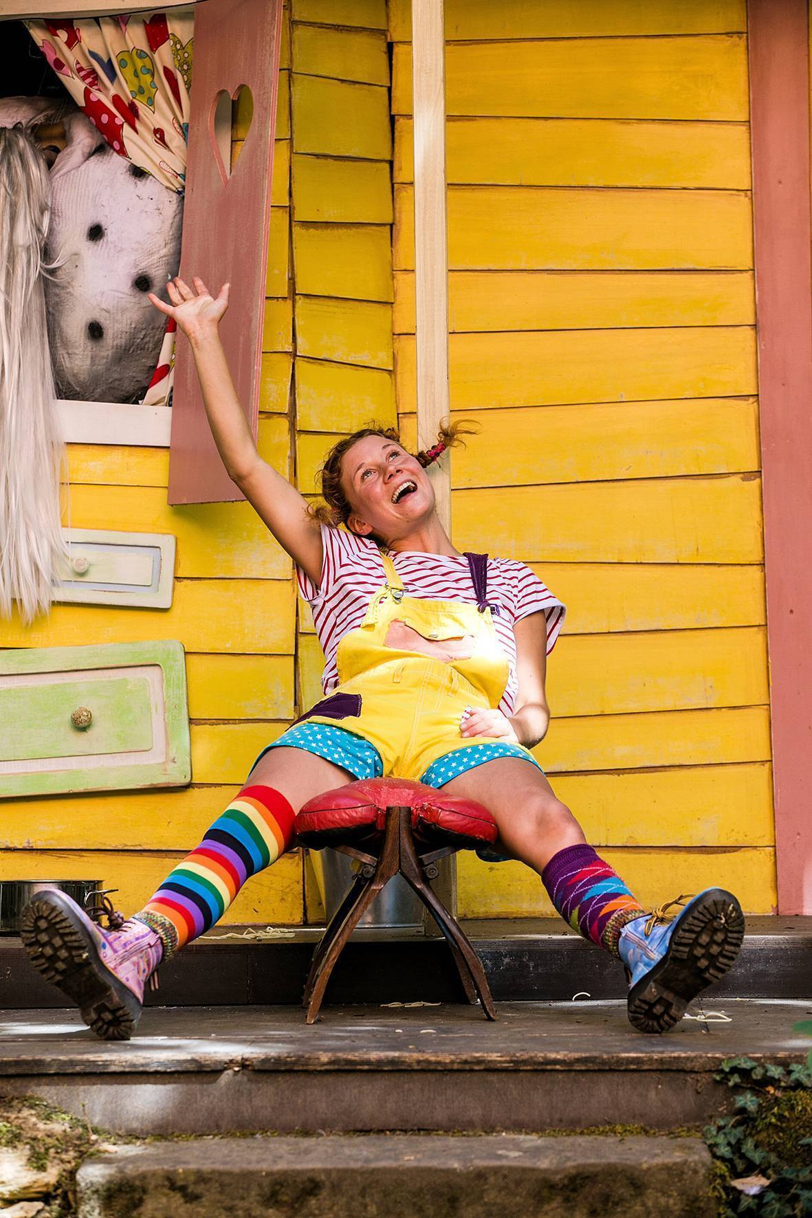 Pippi sitzt fröhlich auf einem Stuhl und reckt den rechten Arm nach oben. Dahinter steht der kleine