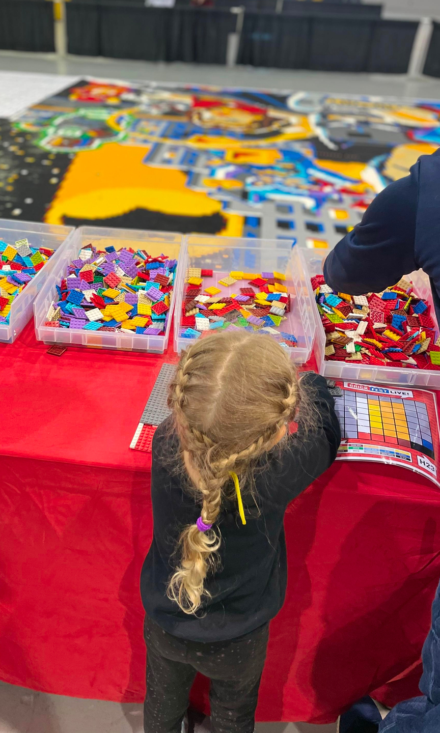 Mädchen steht an einem Tisch, auf dem viele bunte Legosteine in verschiedenen Boxen liegen.