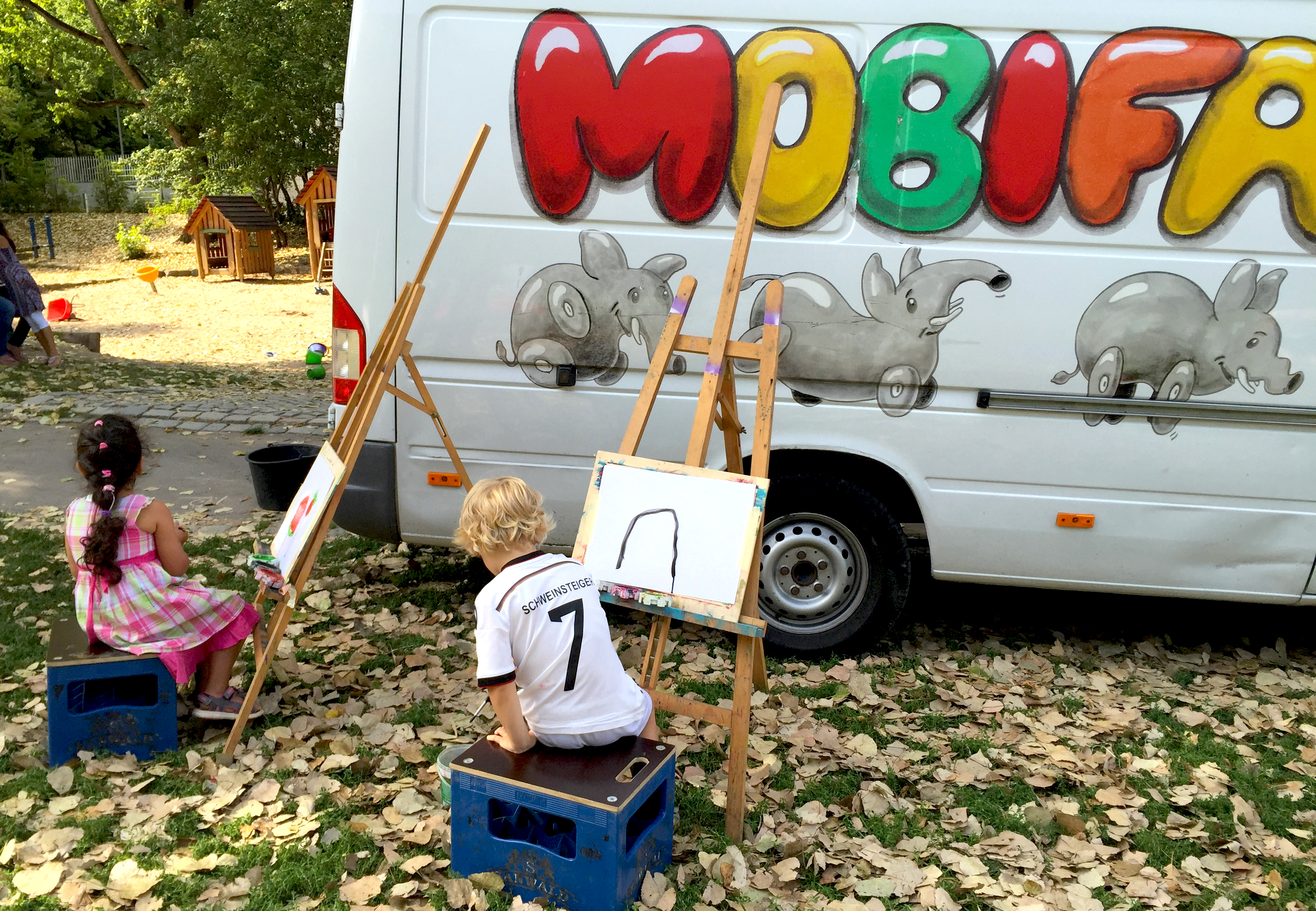 Zwei Kinder sitzen je vor einer Staffelei, dahinter der Mobifant Bus.