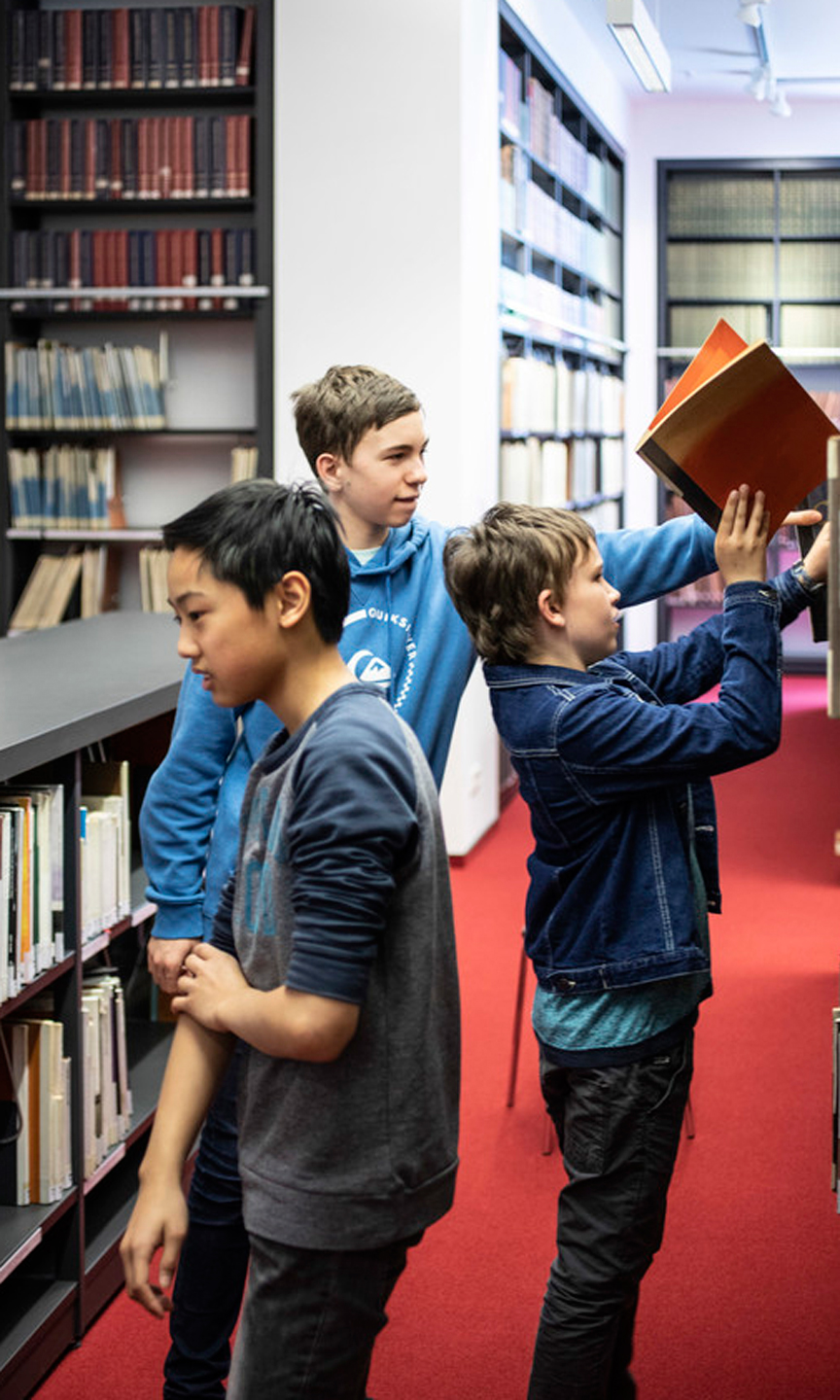 Man sieht drei Jungen zwischen Bücherregalen in einer Bücherei.
