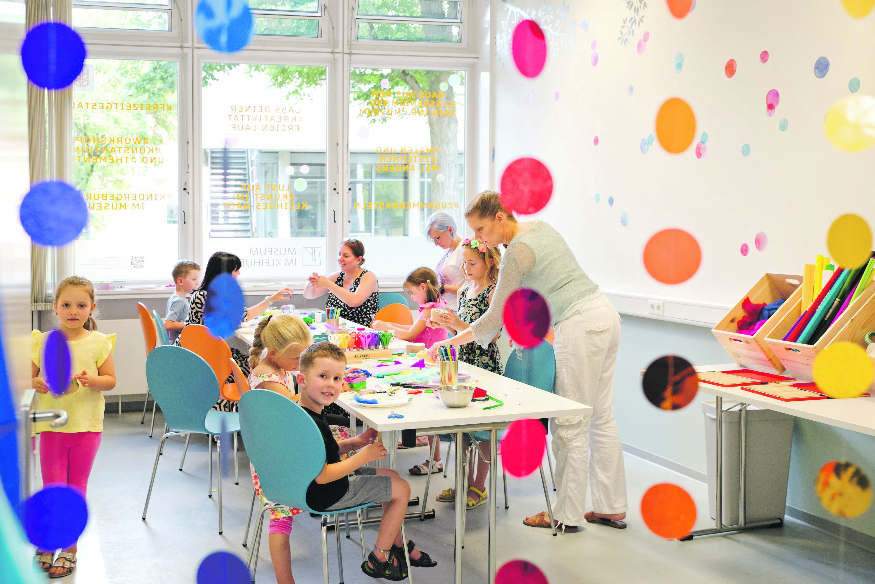 Kinder sitzen an einem Tisch verteilt und Basteln, eine Frau erklärt etwas.