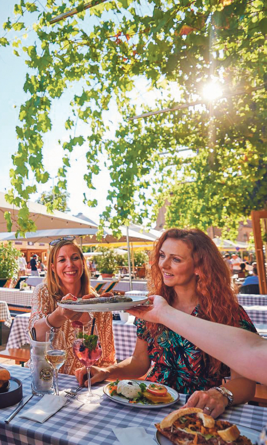 Foto von einem Fest. Unter einem Baum stehen Biergarnituren. Zwei Frauen sitzen da und essen.