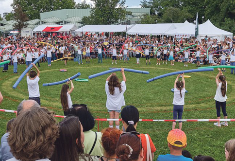 Rasenfläche auf der mehrere Kinder etwas aufführen. Im Kreis stehen andere Menschen drum herum.