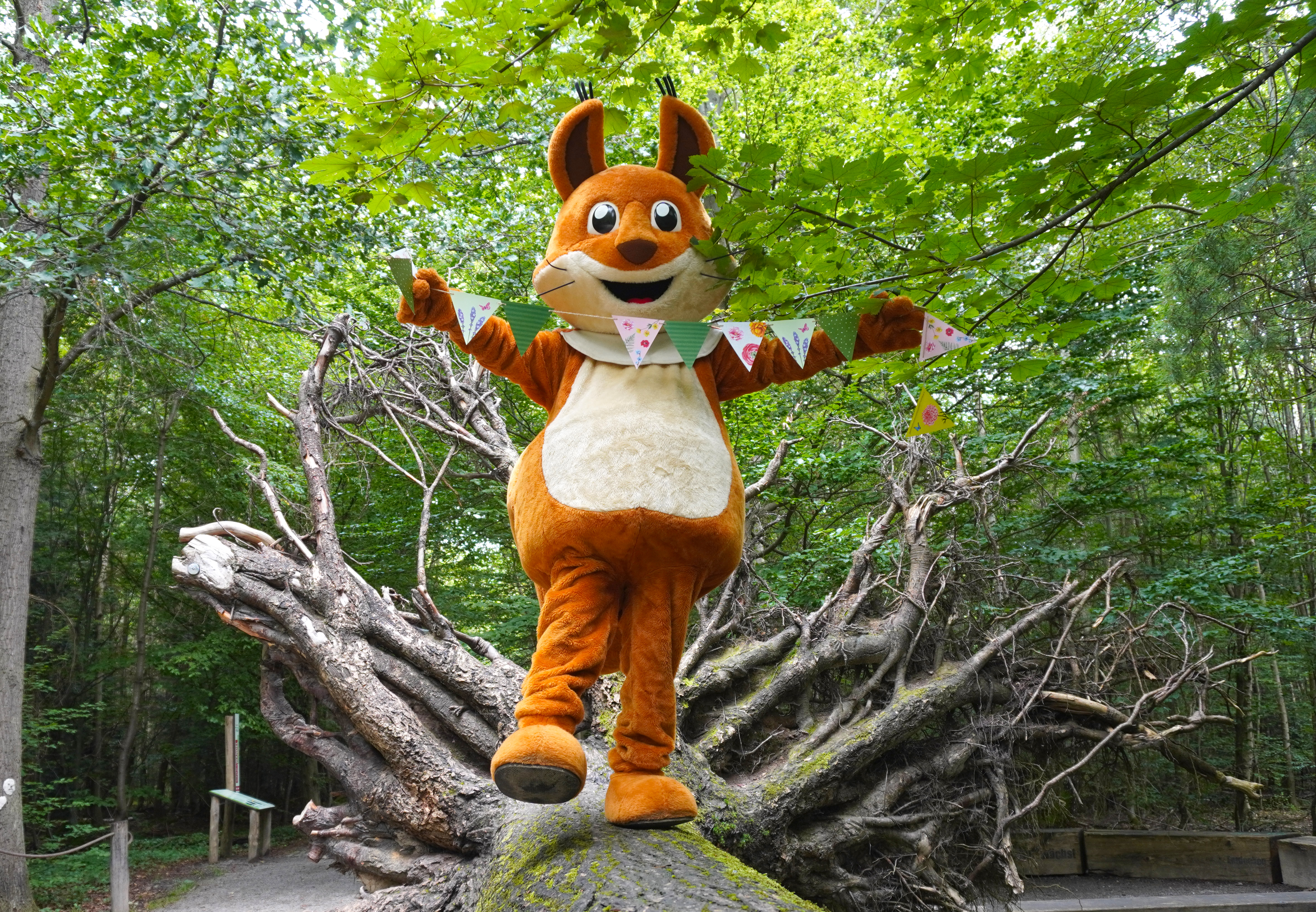 Forsti, das Maskottchen, steht auf einem Baumstamm und hält eine Wimpelkette.