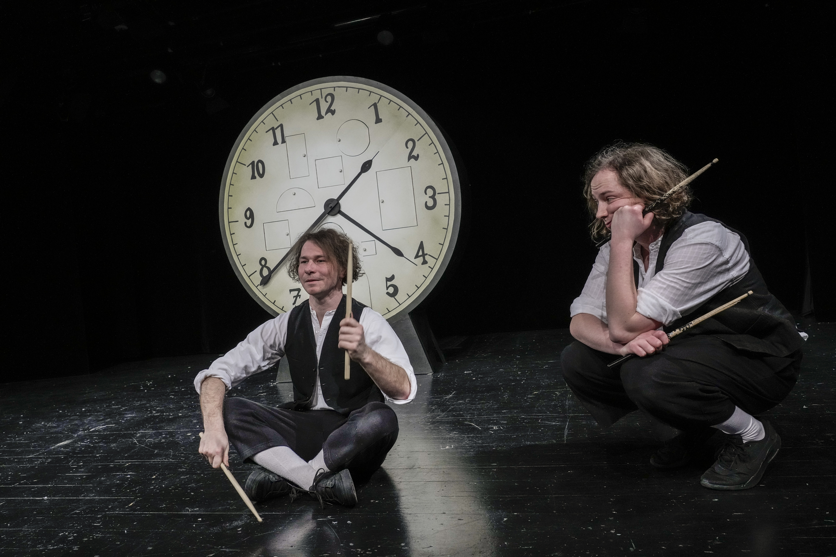 Zwei Männer mit Trommelstöcken sitzen vor einer großen Uhr