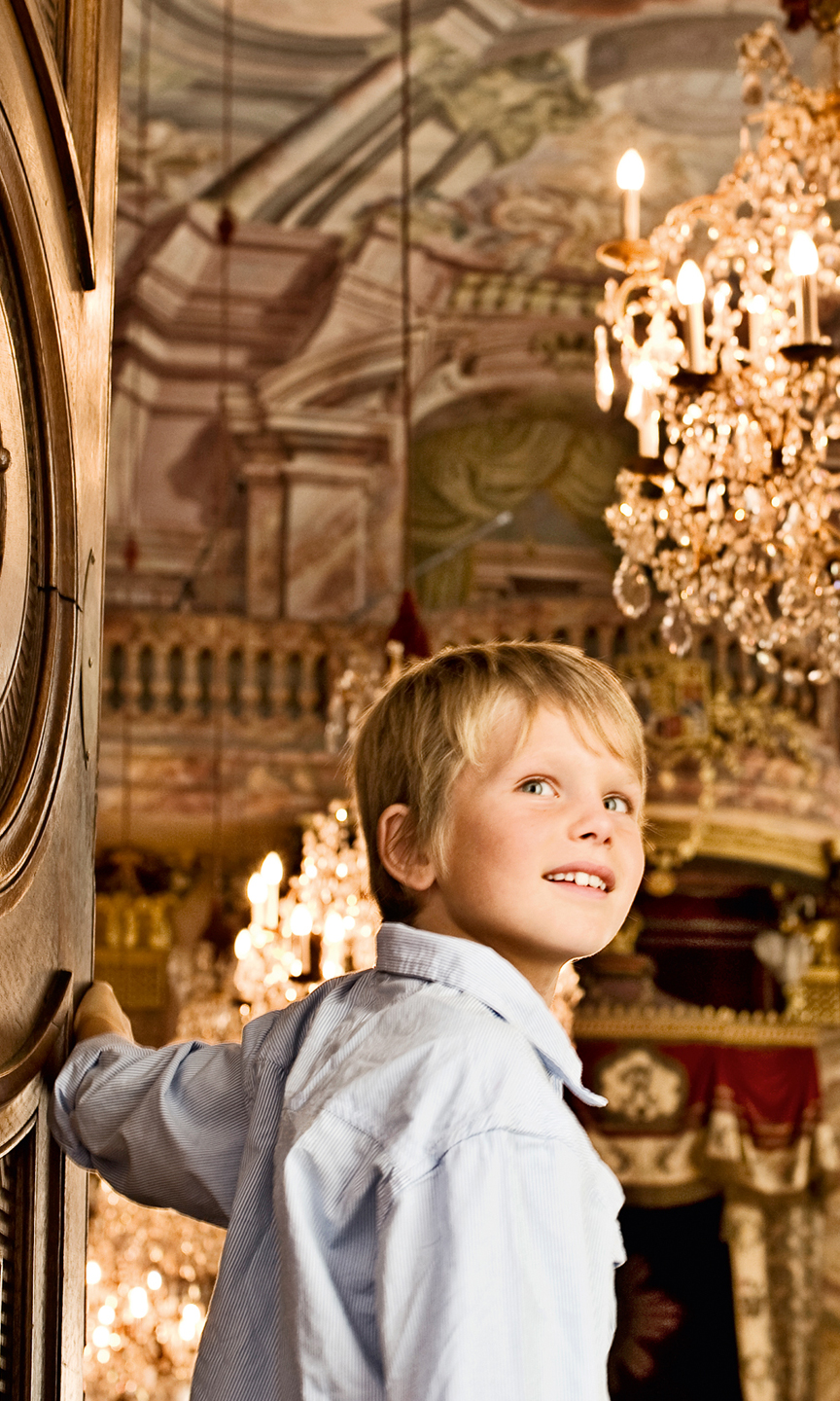 Junge steht in einem Schloss. Er öffnet die Tür zu einem Saal mit Wandmalereien und Kronleuchter.